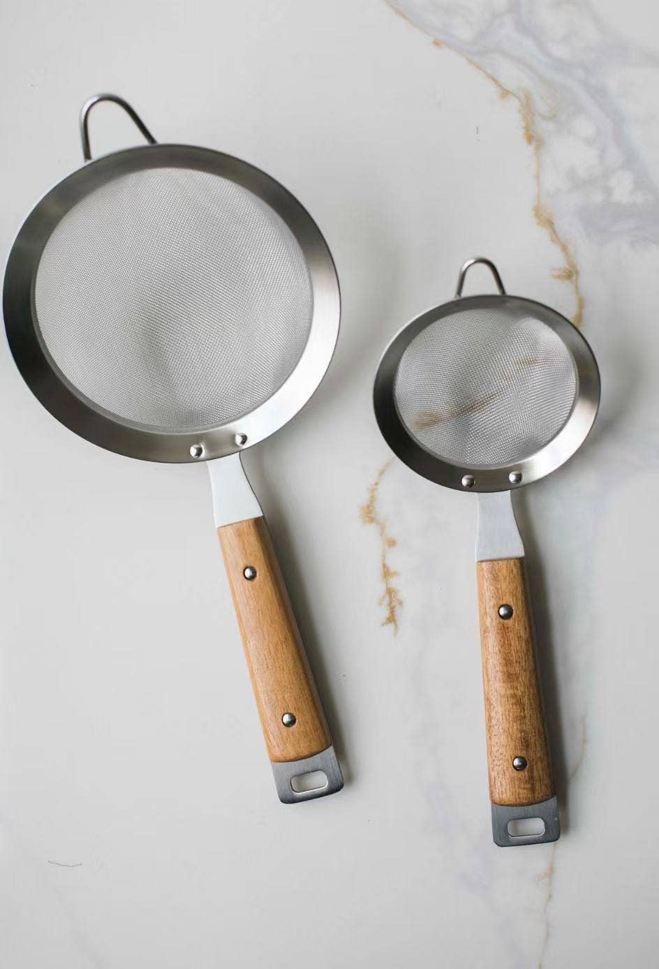 Two metal sieves with wooden handles on a marble surface