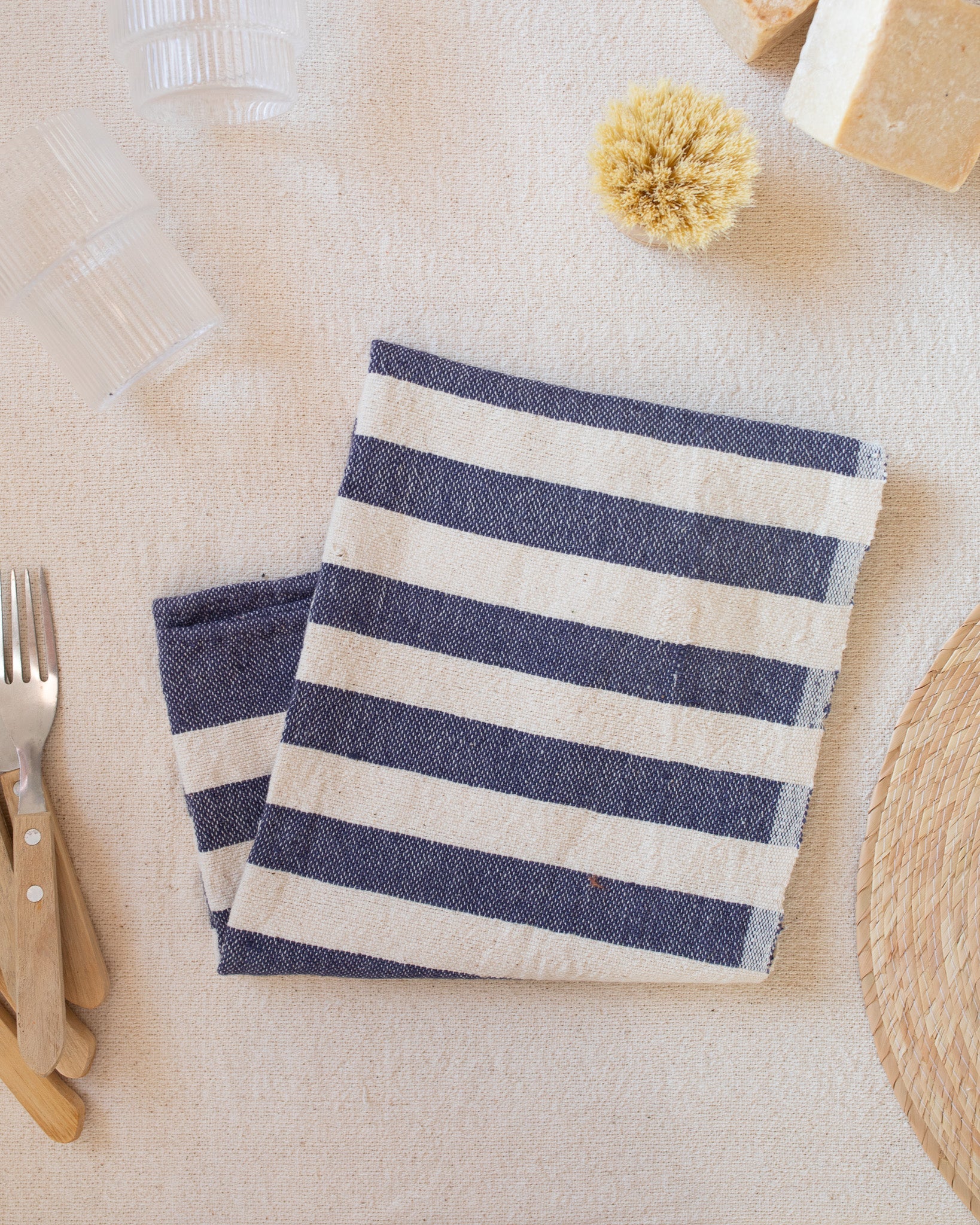 A blue and white striped kitchen towel sitting on a white surface next to utensils and plate.