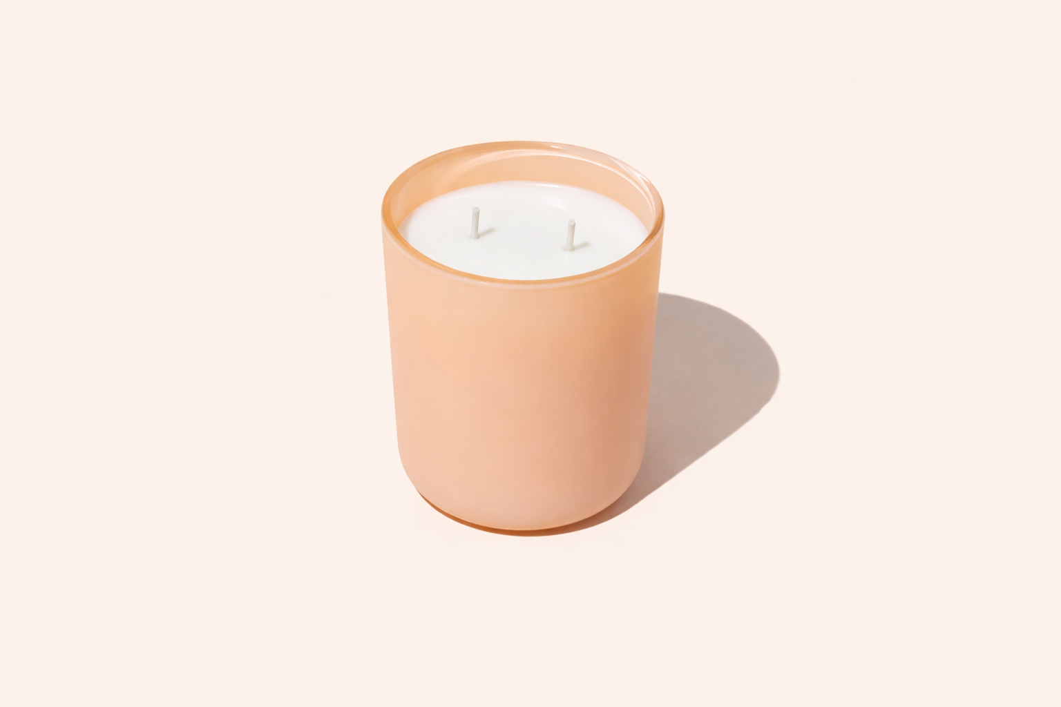 A two-wick scented candle in a frosted peach-colored glass jar with white wax, set against a solid pale beige background with a soft shadow.