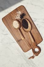 Two Une Vie Nomade walnut wood scoops on a rectangular Une Vie Nomade walnut cutting board, sitting on a white marble surface.