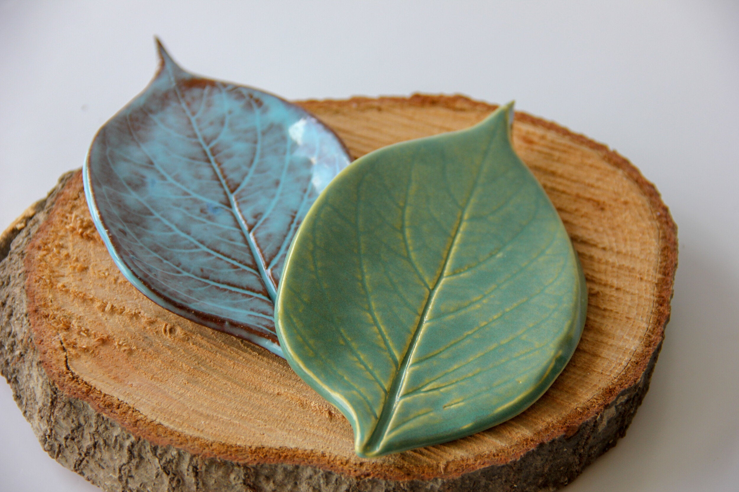 Two green plates in the shape of leaves, sitting on a round piece of wood placed on a white surface.