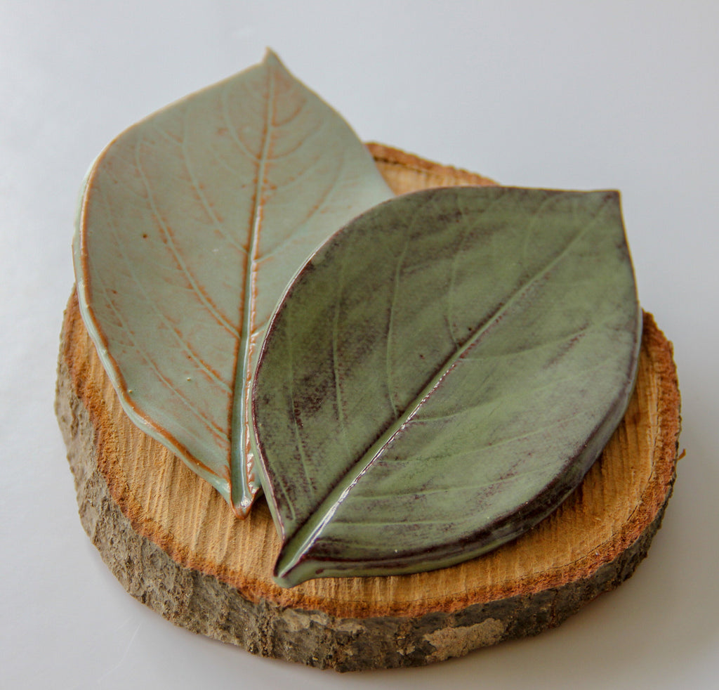 Two green plates in the shape of leaves, sitting on a round piece of wood placed on a white surface.