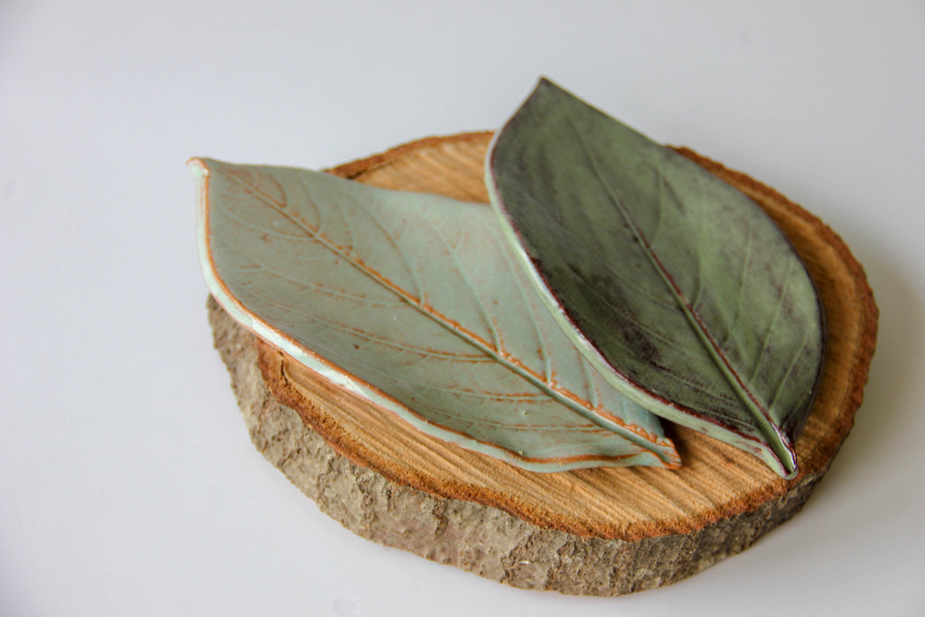 Two green plates in the shape of leaves, sitting on a round piece of wood placed on a white surface.