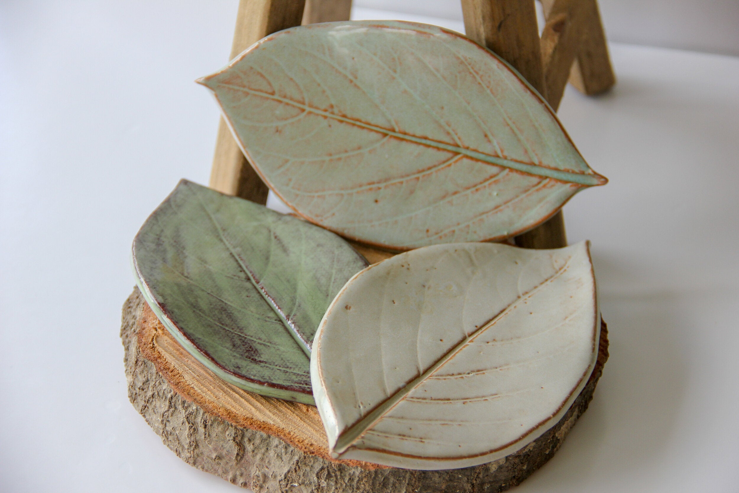 Three green plates in the shape of leaves, sitting on a round piece of wood placed on a white surface.