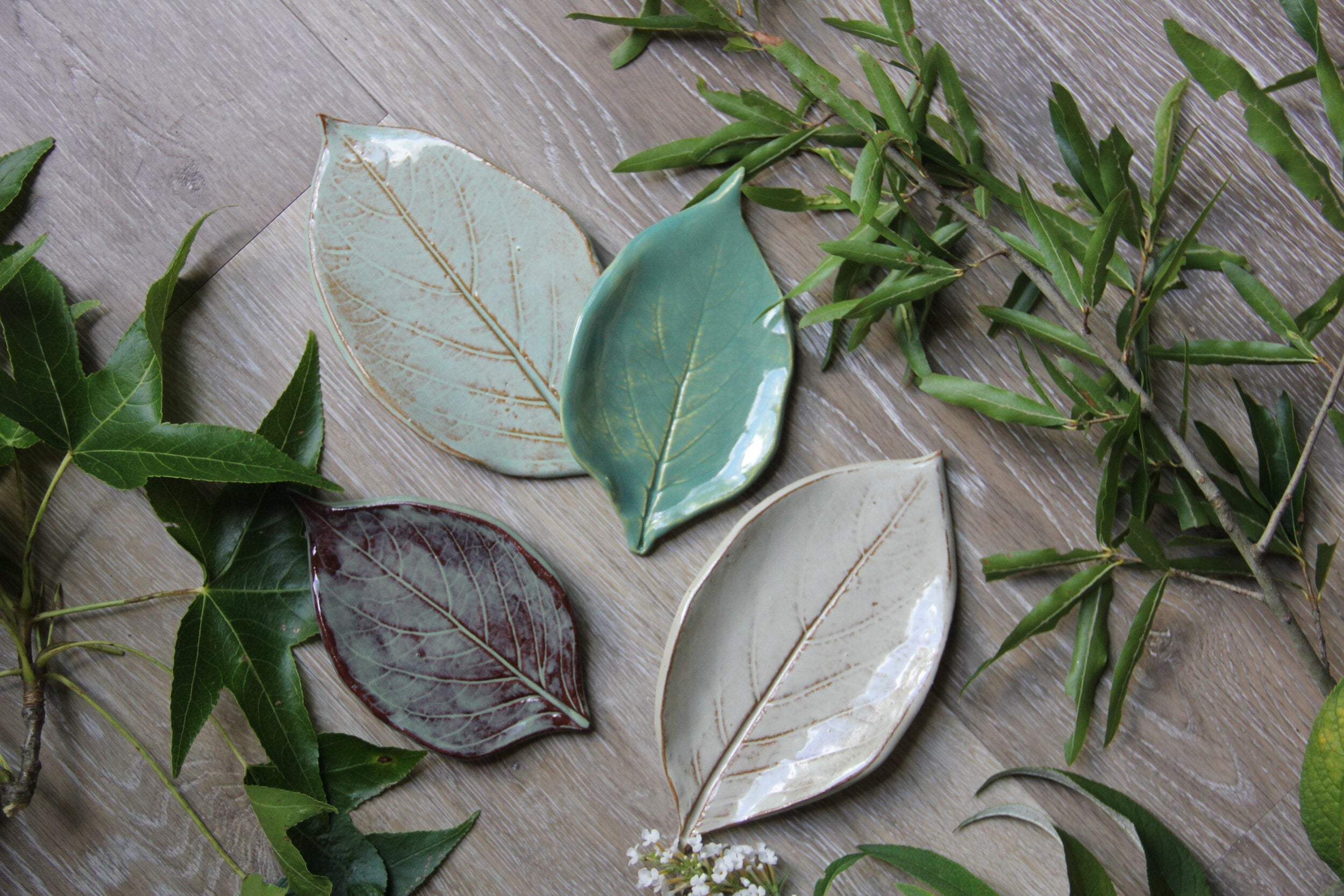 Four green plates in the shape of leaves, sitting on a round piece of wood with greenery placed on a white surface.