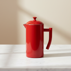 A red Kuchenprofi French press coffee maker with a matching lid and handle, sitting on a white marble countertop against a warm beige background.