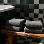 Two stacks of gray towels on a wooden bench in a bathroom with checkered tiles.