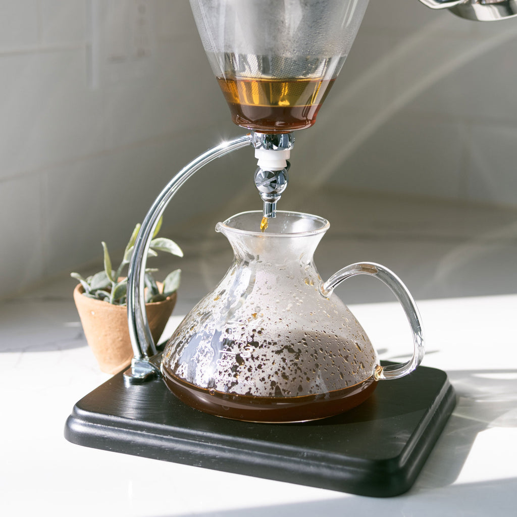 Yama Silverton glass pour over coffee maker with coffee being poured into it on a white surface.